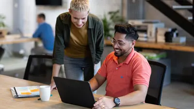Businesswoman standing by male colleague working on laptop at coworking office.