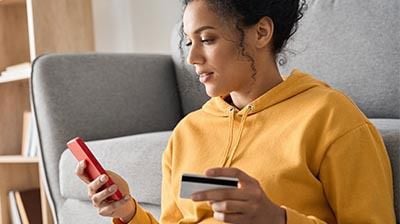A woman in an orange hoodie using a red smart phone while holding a credit card with her other hand