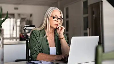 A woman sitting at her kitchen table, using her laptop while resting a hand on her chin 